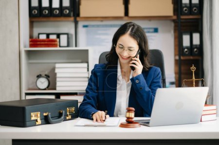 Photo for Asian female lawyer chatting online with client on laptop, discussing details of lawsuit. on smartphone in modern office - Royalty Free Image