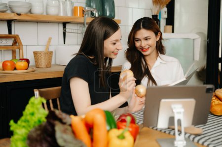 Photo for Two beautiful women in the kitchen in an apron, fresh vegetables on the table, writes down her favourite recipes, comes up with ideas for dishes - Royalty Free Image