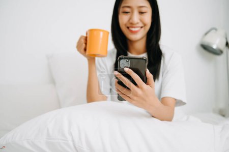 Téléchargez les photos : Belle femme s'est réveillée sur un lit blanc et tenant smartphone et tasse de café le matin à la maison, concept de style de vie - en image libre de droit