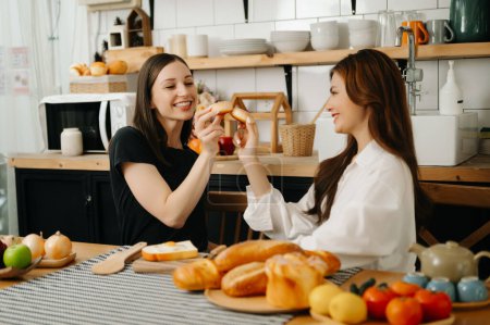 Photo for Two beautiful women in the kitchen in an apron, fresh vegetables on the table, writes down her favourite recipes, comes up with ideas for dishes - Royalty Free Image