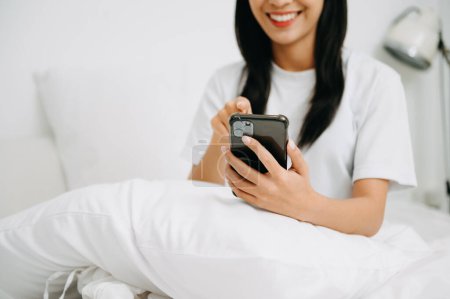 Téléchargez les photos : Belle jeune femme sur un lit blanc en utilisant un smartphone à la maison, concept de style de vie - en image libre de droit