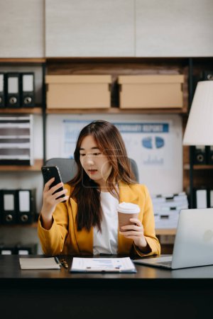 Foto de Confiado hermosa mujer de negocios asiática sosteniendo teléfono inteligente mientras bebe café en la oficina moderna - Imagen libre de derechos