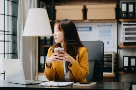 Téléchargez les photos : Confiant belle femme d'affaires asiatique travaillant sur ordinateur portable tout en buvant du café au bureau moderne - en image libre de droit