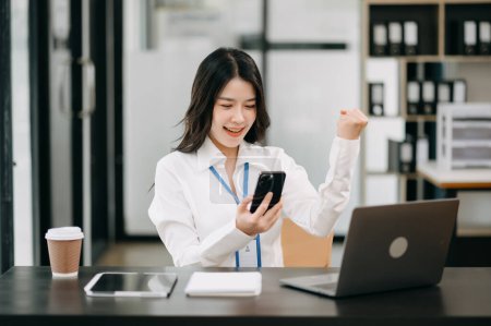 Téléchargez les photos : Heureuse femme d'affaires asiatique au travail au bureau avec smartphone et ordinateur portable - en image libre de droit