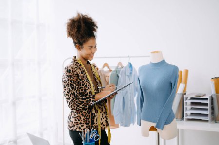 Téléchargez les photos : Femme tailleur africaine travaillant sur des vêtements dans un atelier de couture. belle jeune créatrice de mode sourire et après le succès en studio - en image libre de droit