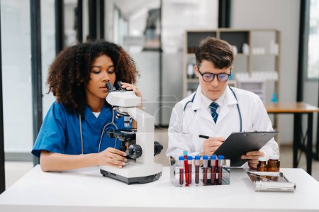Photo for Doctor Talks With Professional Head Nurse or Surgeon, They Use microscope. Diverse Team of Health Care Specialists Discussing Test Result on desk in hospital - Royalty Free Image
