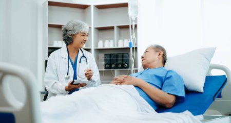 Photo for Asian doctor in white suit talking with elderly patient, who lying on bed in hospital - Royalty Free Image
