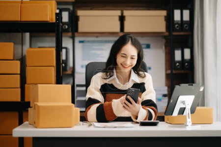 Photo for Startup small business SME,. Asian woman using smartphone, taking receive and checking online purchase shopping order to preparing pack product box. - Royalty Free Image