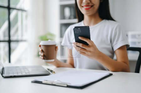 Téléchargez les photos : Confiant belle femme d'affaires asiatique tapant sur smartphone et ordinateur portable tout en tenant le café au bureau - en image libre de droit