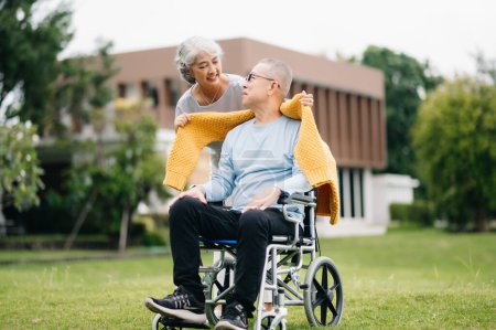 Téléchargez les photos : Asiatique Senior homme assis en fauteuil roulant, femme prenant soin les uns des autres, temps romantique. Ils rient et sourient en plein air dans le parc - en image libre de droit