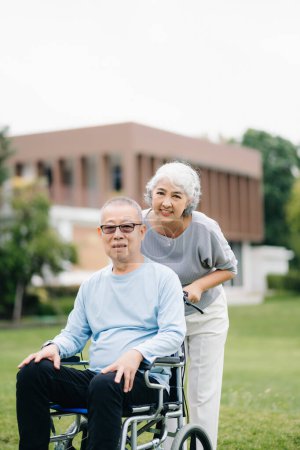 Téléchargez les photos : Asiatique Senior homme assis en fauteuil roulant, femme prenant soin les uns des autres, temps romantique. Ils rient et sourient en plein air dans le parc - en image libre de droit