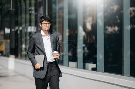 Photo for Handsome male entrepreneur standing outdoors with coffee cup and laptop in hands, leaving office in formal clothes waiting for taxi in street - Royalty Free Image