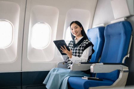 Téléchargez les photos : Attrayant asiatique passager féminin de l'avion assis avec tablette numérique dans un siège confortable. Voyage dans le style - en image libre de droit