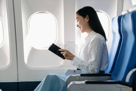 Téléchargez les photos : Attrayant passager asiatique de l'avion tenant carnet, tout en étant assis dans un siège confortable. Voyage dans le style - en image libre de droit