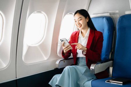 Téléchargez les photos : Attrayant asiatique passager féminin de l'avion assis avec smartphone dans un siège confortable. Voyage dans le style - en image libre de droit