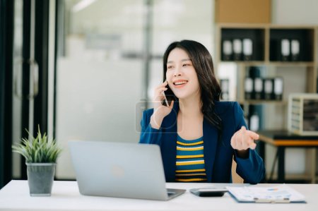 Photo for Smiling Asian woman talking on the phone with a customer. Young positive female accountant using smartphone talking to team at her desk - Royalty Free Image