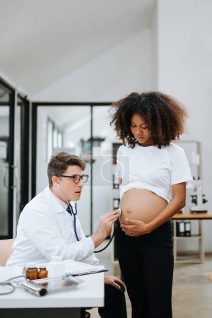 Photo for Pregnant African woman has appointment with doctor at clinic. Male specialist with stethoscope listens to baby's heartbeat in mother's belly. Pregnancy, health care concept - Royalty Free Image