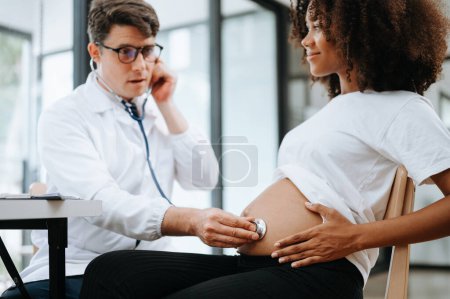 Téléchargez les photos : Femme africaine enceinte a rendez-vous avec le médecin à la clinique. Spécialiste masculin avec stéthoscope écoute le rythme cardiaque du bébé dans le ventre de la mère. Grossesse, concept de soins de santé - en image libre de droit