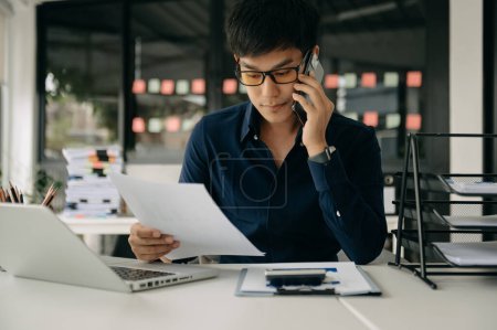 Téléchargez les photos : Jeune homme d'affaires asiatique travaillant au bureau avec ordinateur portable et prenant des notes sur le papier - en image libre de droit