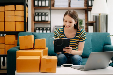 Photo for Young business owner woman prepare parcel box and check online orders for deliver to customer on digital tablet. Shopping Online concept. on sofa at home office - Royalty Free Image