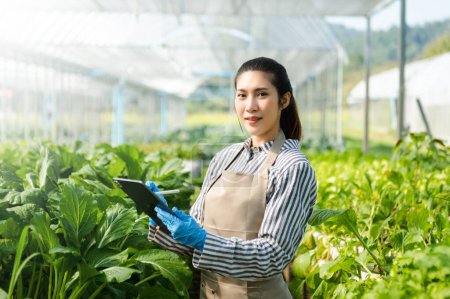 Asian young female farmer in gloves looking at camera using digital tablet in greenhouse plantation. Smart farming technology in agricultural growing activity.