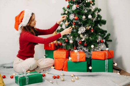 Téléchargez les photos : Femme décorant arbre de Noël avec des jouets. La lumière du soleil inonde le salon. - en image libre de droit