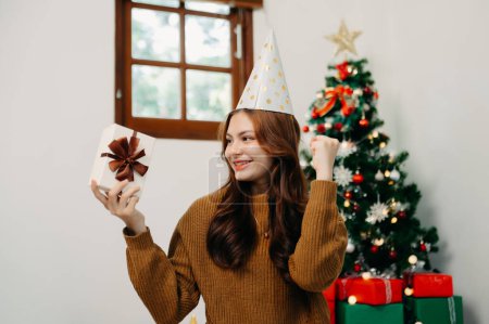 Photo for Asian young happy girl holding present box decorate Christmas tree at home - Royalty Free Image