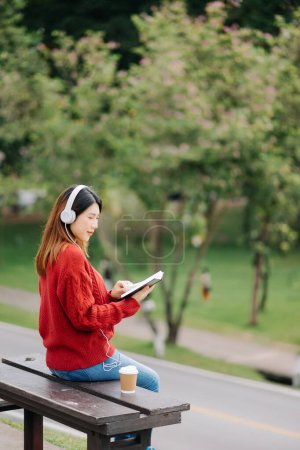 Téléchargez les photos : Belle jeune femme asiatique écoutant de la musique avec des écouteurs avec le sentiment heureux et détendu, frais avec le visage souriant. dans le parc le matin - en image libre de droit
