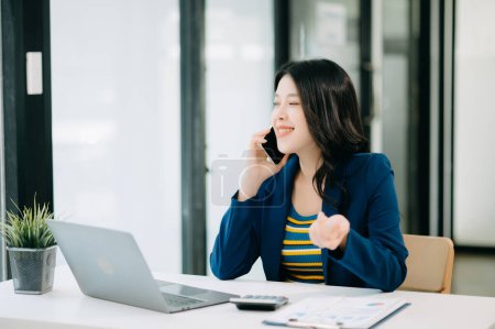 Photo for Smiling Asian woman talking on the phone with a customer. Young positive female accountant using smartphone talking to team at her desk - Royalty Free Image