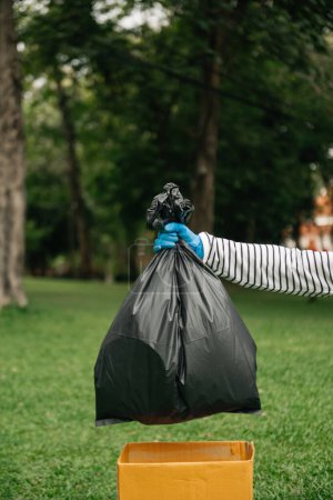 Foto de Mano sosteniendo la basura bolsa negra poner en la basura para limpiar. Aclaración, contaminación, ecología y concepto plástico. en el parque - Imagen libre de derechos