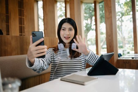 Téléchargez les photos : Attrayant jeune étudiant asiatique heureux étudiant à la bibliothèque du collège, assis au bureau, en utilisant un smartphone, tablette numérique et écouteurs ayant un chat vidéo - en image libre de droit