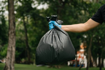 Foto de Mano sosteniendo la basura bolsa negra poner en la basura para limpiar. Aclaración, contaminación, ecología y concepto plástico en el parque - Imagen libre de derechos