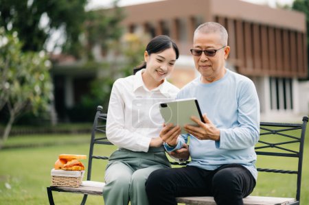 Elderly asian senior man on wheelchair with Asian careful caregiver with tablet pc and food in hospital garden concept.