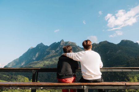 Téléchargez les photos : Vue arrière de deux voyageurs masculins assis et regardant une belle vue sur la montagne, le ciel et la nature dans la lumière du matin - en image libre de droit