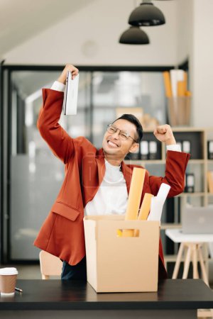 Téléchargez les photos : Joyeux et excité jeune Asiatique employé de bureau célébrant sa démission, portant ses affaires personnelles. quitter un emploi, changer ou une entreprise dans un bureau moderne - en image libre de droit