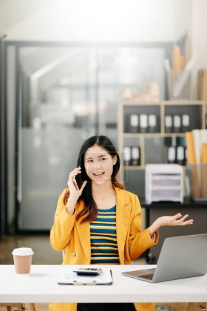 Foto de Mujer de negocios asiática Hablando en el teléfono inteligente y utilizando un ordenador portátil con una sonrisa mientras está sentado en la oficina moderna - Imagen libre de derechos