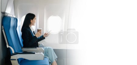 Foto de Atractiva pasajera asiática del avión sentada en un asiento cómodo mientras trabaja en un smartphone con área simulada usando conexión inalámbrica. Viaja con estilo, trabaja con gracia - Imagen libre de derechos
