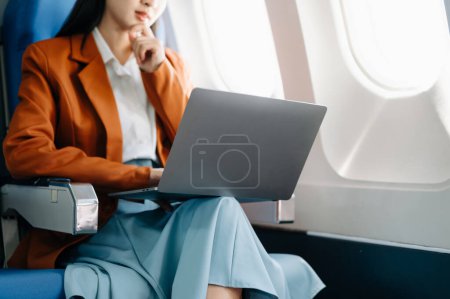 Foto de Atractiva pasajera asiática del avión sentada en un asiento cómodo mientras trabaja en una computadora portátil con área simulada usando conexión inalámbrica. Viaja con estilo, trabaja con gracia - Imagen libre de derechos