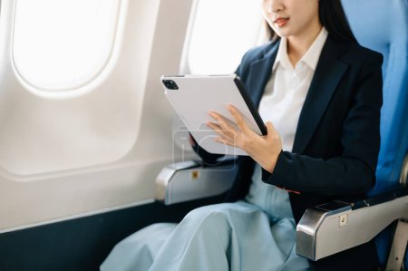 Foto de Atractiva pasajera asiática de avión sentada en asiento cómodo mientras trabaja en tableta digital con área de maqueta utilizando conexión inalámbrica. Viaja con estilo, trabaja con gracia - Imagen libre de derechos