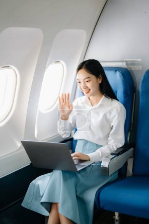 Téléchargez les photos : Attrayant passager asiatique féminine de l'avion assis dans un siège confortable tout en travaillant sur un ordinateur portable avec zone maquette en utilisant une connexion sans fil. Voyagez avec style, travaillez avec grâce - en image libre de droit