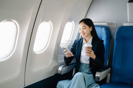 Téléchargez les photos : Attrayant passager asiatique de l'avion assis dans un siège confortable tout en travaillant sur smartphone avec zone maquette en utilisant une connexion sans fil. Voyagez avec style, travaillez avec grâce - en image libre de droit