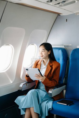 Photo for Attractive Asian female passenger of airplane sitting in comfortable seat while working on digital tablet with mock up area using wireless connection. Travel in style, work with grace - Royalty Free Image