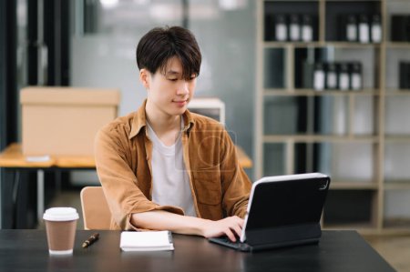 Téléchargez les photos : Homme asiatique confiant avec un sourire travaillant sur tablette informatique au bureau moderne - en image libre de droit