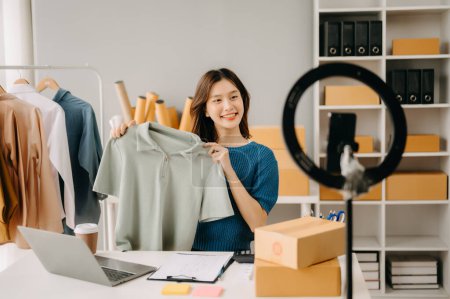 Foto de Concepto de blogger de moda, Mujer joven asiática vendiendo ropa en streaming de video. Puesta en marcha de pequeñas empresas PYME, utilizando smartphone - Imagen libre de derechos