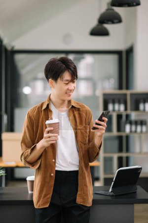 Téléchargez les photos : Homme d'affaires asiatique confiant tapant sur smartphone tout en tenant un café au bureau - en image libre de droit
