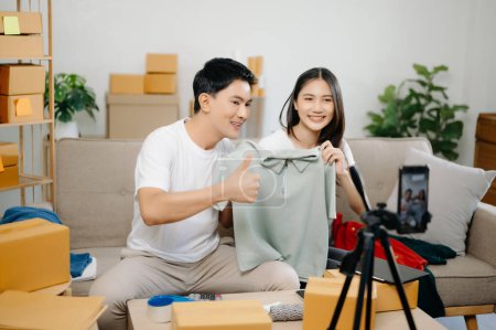 Foto de Dos bloggers asiáticos mostrando ropa frente a la cámara para grabar vídeo vlog en directo en su tienda. Concepción de carrito de compras en línea. en el sofá en casa - Imagen libre de derechos