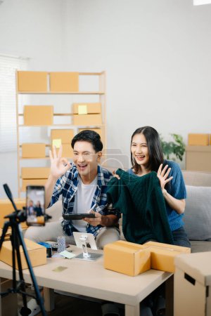 Photo for Two Asian bloggers showing clothes in front of the camera to recording vlog video live streaming at her shop. Online Shopping Cart notion. on sofa at home - Royalty Free Image