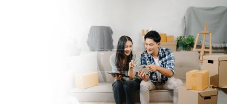 Photo for Asian young attractive couple man and woman use tablet and smartphone for online shopping furniture, decorate house, carton package move in new house. Young married asian moving home. - Royalty Free Image
