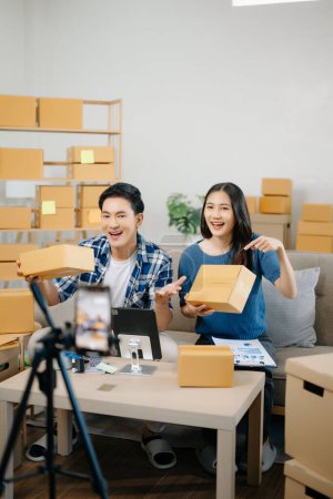 Photo for Two Asian bloggers showing clothes in front of the camera to recording vlog video live streaming at her shop. Online Shopping Cart notion. on sofa at home - Royalty Free Image