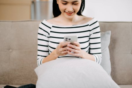 Foto de Joven mujer asiática relajarse en cómodo sofá en casa mensajes de texto de mensajería en el teléfono inteligente, chica sonriente utilizar las compras de teléfonos celulares en línea desde casa en el sofá - Imagen libre de derechos